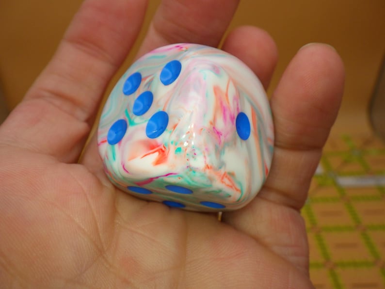 May include: A six-sided die with a marbled, multi-colored surface featuring swirls of white, pink, and teal. The die has rounded edges and is marked with bright blue circular pips on its faces, showing the numbers three and one clearly.