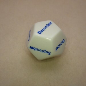 Pode incluir: Dado branco de seis lados com letras azuis. As faces mostram os meses de setembro, outubro e dezembro.