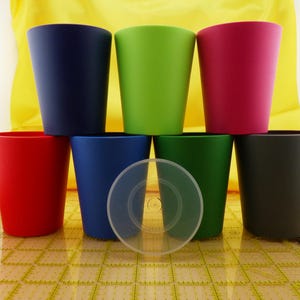 May include: A collection of eight colorful tumblers in shades of red, blue, green, and pink. A clear plastic lid rests in front of the tumblers. The tumblers are arranged in two rows, with a yellow background.