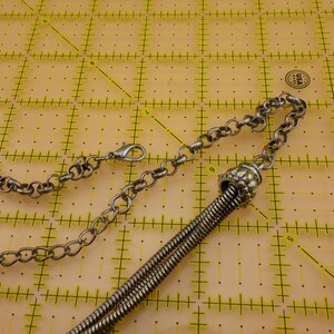 May include: A silver-toned metal chain necklace featuring a lobster claw clasp and a decorative textured cap that secures a series of thin, flexible metal strands. The necklace is positioned on a yellow cutting mat with visible inch markings.