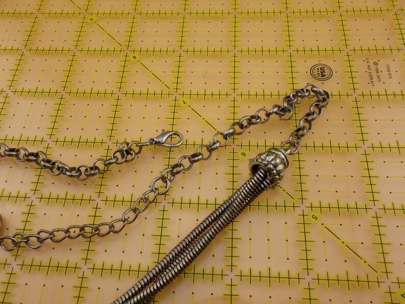 May include: A silver-toned metal chain necklace featuring a lobster claw clasp and a decorative textured cap that secures a series of thin, flexible metal strands. The necklace is positioned on a yellow cutting mat with visible inch markings.
