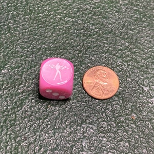 May include: A pink dice with a white silhouette of a person on one side and white dots on the other sides. The dice is next to a copper penny.