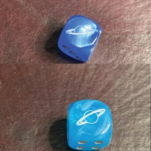 May include: Two blue dice with white swirl patterns and a white planet with rings design on each face. The dice are on a brown surface.