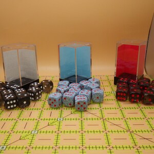 May include: An assortment of dice in various colors and styles. There are black dice with white dots, blue dice with red dots, and red dice with black dots. The dice are arranged in front of clear, blue, and red acrylic boxes.