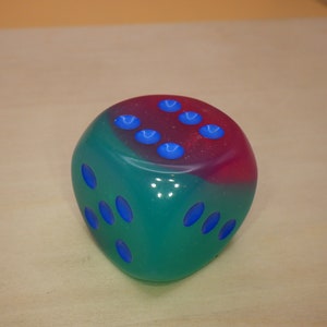 May include: A two-toned green and pink dice with blue dots. The dice is made of a translucent material.