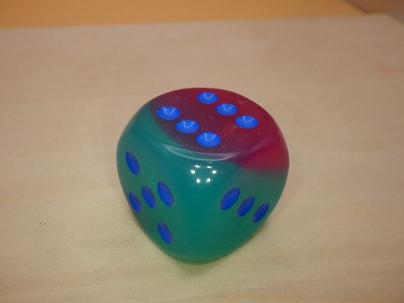 May include: A two-toned green and pink dice with blue dots. The dice is made of a translucent material.