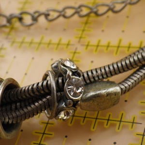 May include: A silver-toned snake chain bracelet featuring a central charm embedded with clear, round-cut crystals. The bracelet is positioned over a yellow measuring mat with markings in inches, showing the chain width is approximately 0.25 inches.