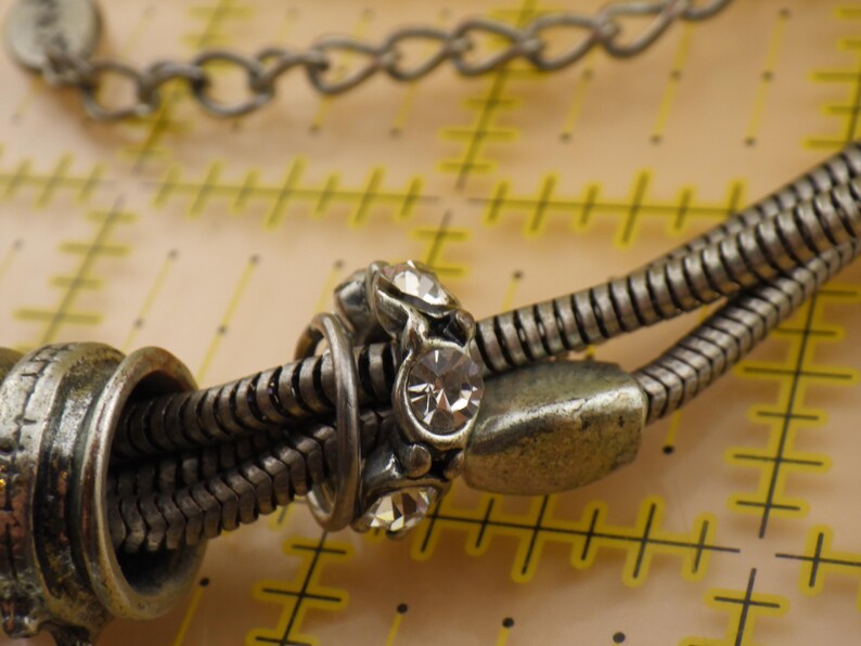 May include: A silver-toned snake chain bracelet featuring a central charm embedded with clear, round-cut crystals. The bracelet is positioned over a yellow measuring mat with markings in inches, showing the chain width is approximately 0.25 inches.