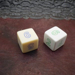 May include: Two dice, one cream-colored and one white, are displayed. The cream die shows the numbers 5 and 6 in blue, while the white die displays 2, 4, and 6 in green. The dice are on a dark, textured surface.