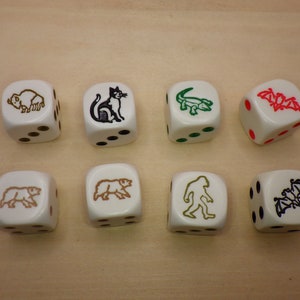 May include: Eight white dice with different animal illustrations. The dice are on a light brown wooden surface. The animals include a bison, a cat, an alligator, a bat, a bear, a caveman, and two bats.