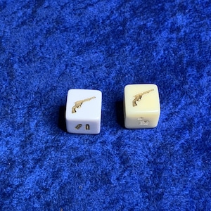 May include: Two white dice with gold accents. One die has a picture of a gun and two bullets. The other die has a picture of a gun and two bullets.