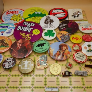 May include: A collection of vintage buttons and pins with various designs and text. The assortment includes a hot air balloon, a frog, a shamrock, and the text "Smile God Loves You". There are also pins with the text "American" and "Watch Your Step".