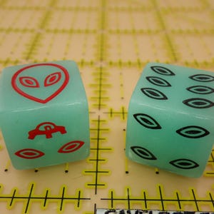 May include: Two green dice with different designs. One die has a red alien face and a red flying saucer. The other die has six black eye shapes.