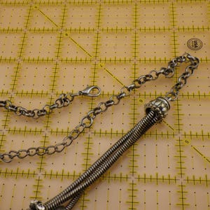May include: A silver-toned metal necklace featuring a thick, coiled snake chain segment connected to a standard round link chain. The necklace includes a lobster claw clasp, an adjustable extender chain, and a small oval tag with engraved text.