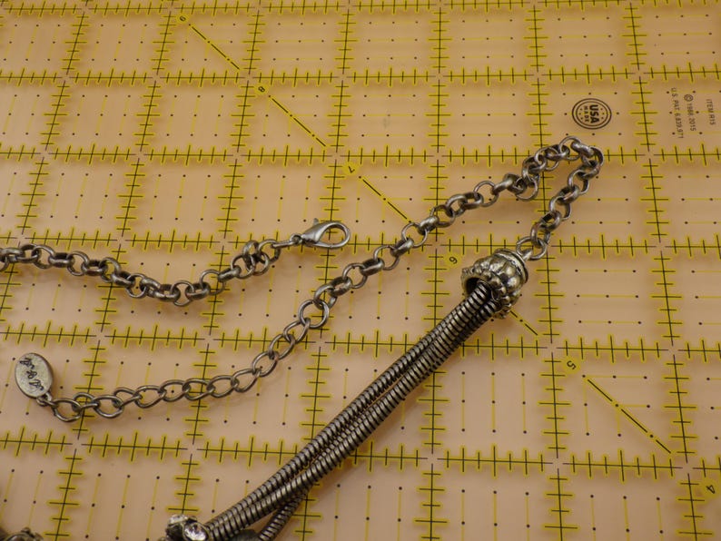 May include: A silver-toned metal necklace featuring a thick, coiled snake chain segment connected to a standard round link chain. The necklace includes a lobster claw clasp, an adjustable extender chain, and a small oval tag with engraved text.