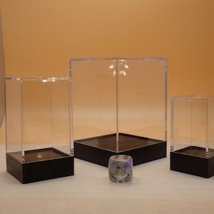 May include: Three clear acrylic display boxes with black bases. The boxes are different sizes and are arranged in a row on a white surface. A small, clear, multi-colored dice is in front of the boxes.