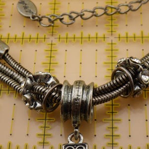 May include: A silver-toned multi-strand snake chain necklace featuring decorative metal beads and clear rhinestones. The necklace includes a central cylindrical bead and a small metal tag on the chain, positioned over a yellow measuring mat.