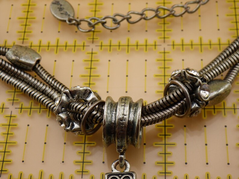 May include: A silver-toned multi-strand snake chain necklace featuring decorative metal beads and clear rhinestones. The necklace includes a central cylindrical bead and a small metal tag on the chain, positioned over a yellow measuring mat.
