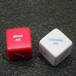 May include: Two dice with text. One red die reads "BROAD AXE" and "HAND AXE". The other die is white and reads "THROWING AXE". The dice are on a textured, dark surface.