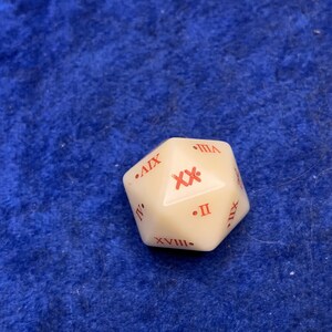 May include: Ivory-colored twenty-sided die with red Roman numerals. The die is a dodecahedron shape, with each face displaying a different numeral. The die is set against a blue background.