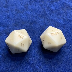 May include: Two ivory-colored, multi-sided dice with Roman numerals etched on each face. The dice are displayed on a blue textured surface. The dice are likely used for gaming or collecting.