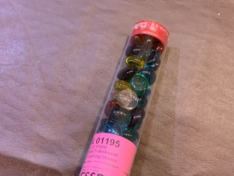 May include: A clear plastic tube filled with colorful glass gaming stones. The tube has a pink label with the text "K 01195 1/2 or more Red Translucent Gaming Stones".