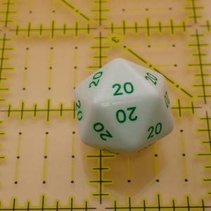 May include: A white twenty-sided die featuring green numbers. The number 20 is printed on multiple visible faces of the die. The die is positioned on a yellow cutting mat with black grid lines and markings in inches for scale reference.