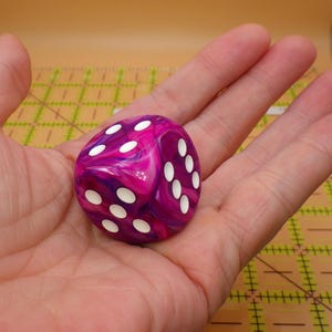 May include: A six-sided die with a marbled pattern of deep magenta, purple, and white swirls. The die features rounded corners and recessed white circular pips on its faces, showing the numbers three, four, and five clearly in this perspective.