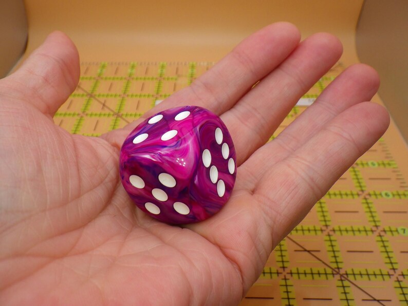 May include: A six-sided die with a marbled pattern of deep magenta, purple, and white swirls. The die features rounded corners and recessed white circular pips on its faces, showing the numbers three, four, and five clearly in this perspective.