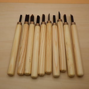 May include: A set of ten wood carving tools with light-colored wooden handles and dark metal blades. The tools are arranged in a row, showcasing their various blade shapes and sizes. The handles have a smooth, natural finish, and the blades appear sharp.