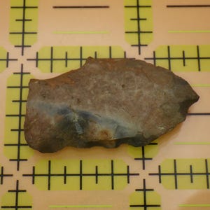 May include: A roughly shaped, brown stone tool with a lighter, blue-grey streak. The stone is set against a yellow background with black grid lines, likely for measurement.