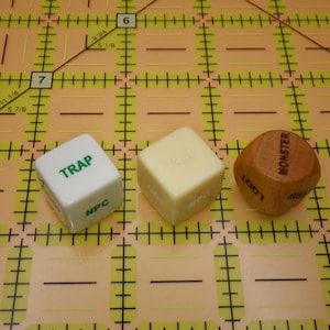 May include: Three six-sided dice arranged in a row on a yellow grid mat. The first die is white with green text reading TRAP and NPC. The second die is cream-colored with faint text. The third die is brown wood with black text reading MONSTER and LOOT.