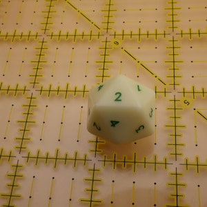 May include: An off-white twenty-sided die featuring green numerical markings. The die is positioned on a translucent yellow cutting mat that displays a grid of black lines and markings, providing a clear visual reference for the object's scale.