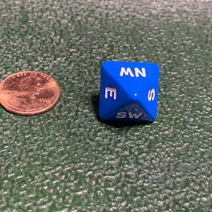 May include: A blue six-sided die with white letters showing the directions North, South, East, West, Northwest, and Southwest.
