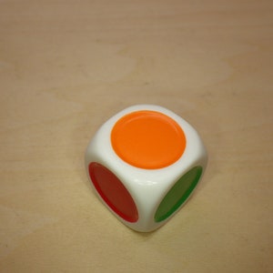 May include: A white cube with three colored circles on its sides. The top circle is orange, the side circle is red, and the bottom circle is green.