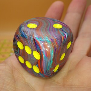 May include: A six-sided die featuring a swirling, multi-colored marbled pattern of blue, purple, orange, and teal. The die has rounded corners and bright neon yellow circular pips recessed into the surface on the visible faces of the cube.