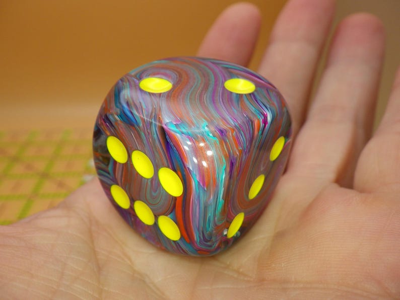 May include: A six-sided die featuring a swirling, multi-colored marbled pattern of blue, purple, orange, and teal. The die has rounded corners and bright neon yellow circular pips recessed into the surface on the visible faces of the cube.