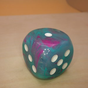 May include: A teal and pink swirled resin dice with white pips. The dice is a six-sided cube.