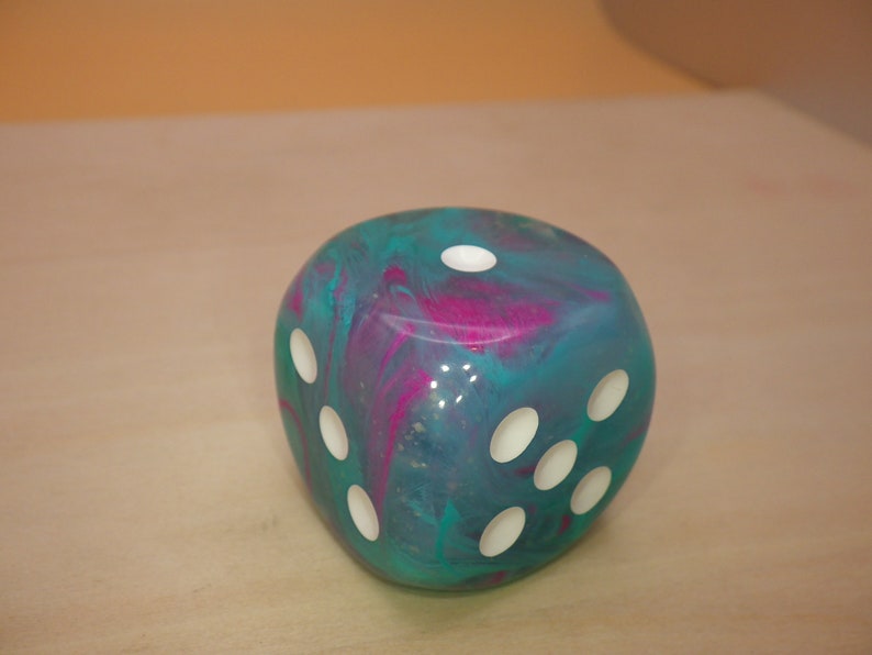 May include: A teal and pink swirled resin dice with white pips. The dice is a six-sided cube.