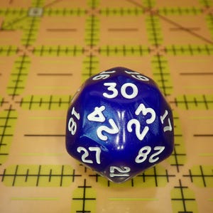 May include: A dark blue, multi-faceted polyhedral die featuring white engraved numbers. The die displays the number 30 at the top, with surrounding numbers including 24, 23, 28, 27, 21, 18, and 17 clearly visible on its various faces.