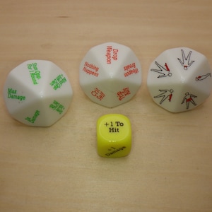 May include: Three white multi-sided dice with printed text and illustrations, alongside one small yellow six-sided die. The white dice feature red, green, and black text including phrases like Max Damage, Drop Weapon, and Crit Self, plus human figure icons.