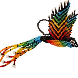 May include: A handcrafted, beaded hummingbird in flight. The bird features a vibrant array of colors, including blue, green, yellow, orange, and red, with black accents. The wings are spread, and the tail feathers are long and flowing.