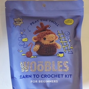 May include: A blue package featuring a crochet kit for beginners. The package has the text "Peat the Troll" and "The Woobles" in glitter. The kit includes all materials and glitter yarn. A crocheted troll is pictured.