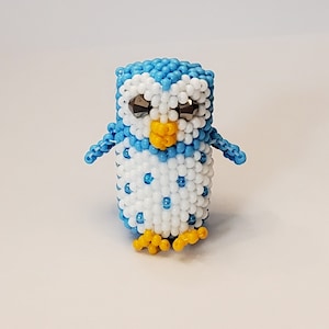 May include: A small blue and white beaded penguin with yellow feet and black eyes. The penguin is made of tiny beads and is a cute and whimsical decoration.