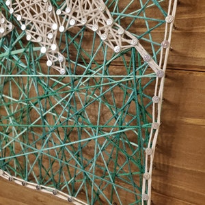 Puede incluir: Una pieza de arte de hilo con un dise&ntilde;o de hilo blanco sobre un fondo de madera. El arte de hilo est&aacute; hecho con hilo verde y azul, creando un efecto degradado.