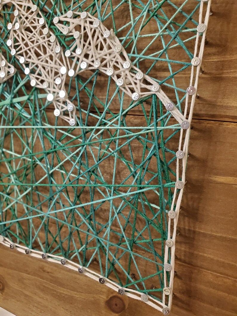 Puede incluir: Una pieza de arte de hilo con un dise&ntilde;o de hilo blanco sobre un fondo de madera. El arte de hilo est&aacute; hecho con hilo verde y azul, creando un efecto degradado.