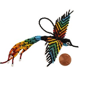 May include: A beaded hummingbird ornament with a rainbow of colors, including red, orange, yellow, green, and blue. The bird is black with a long tail and wings. A penny is next to the hummingbird for size comparison.