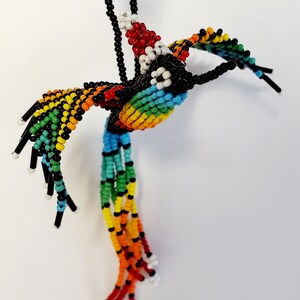 May include: A beaded hummingbird pendant necklace. The bird features a rainbow gradient across its wings and body, with black and white accents. The pendant hangs from a black beaded chain. The hummingbird's tail is made of long, colorful beaded strands.