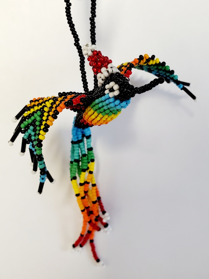 May include: A beaded hummingbird pendant necklace. The bird features a rainbow gradient across its wings and body, with black and white accents. The pendant hangs from a black beaded chain. The hummingbird's tail is made of long, colorful beaded strands.