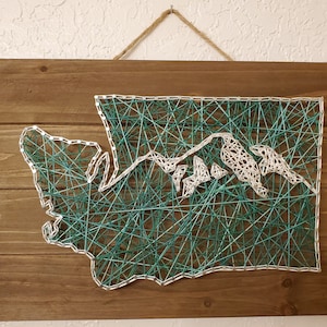 May include: A wooden wall art piece featuring a string art design of the state of Washington. The outline of the state is created with white string, and the mountains are filled in with teal string.